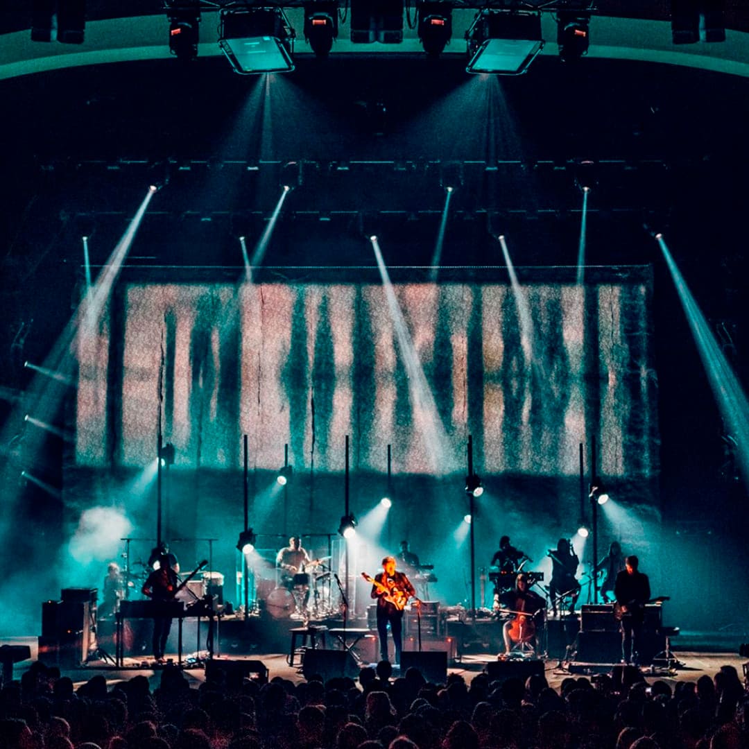 La imagen muestra una banda tocando en vivo en un escenario iluminado con luces azules. Los músicos tocan guitarra, batería y otros instrumentos, mientras una gran pantalla detrás proyecta imágenes abstractas, creando una atmósfera visual impactante.