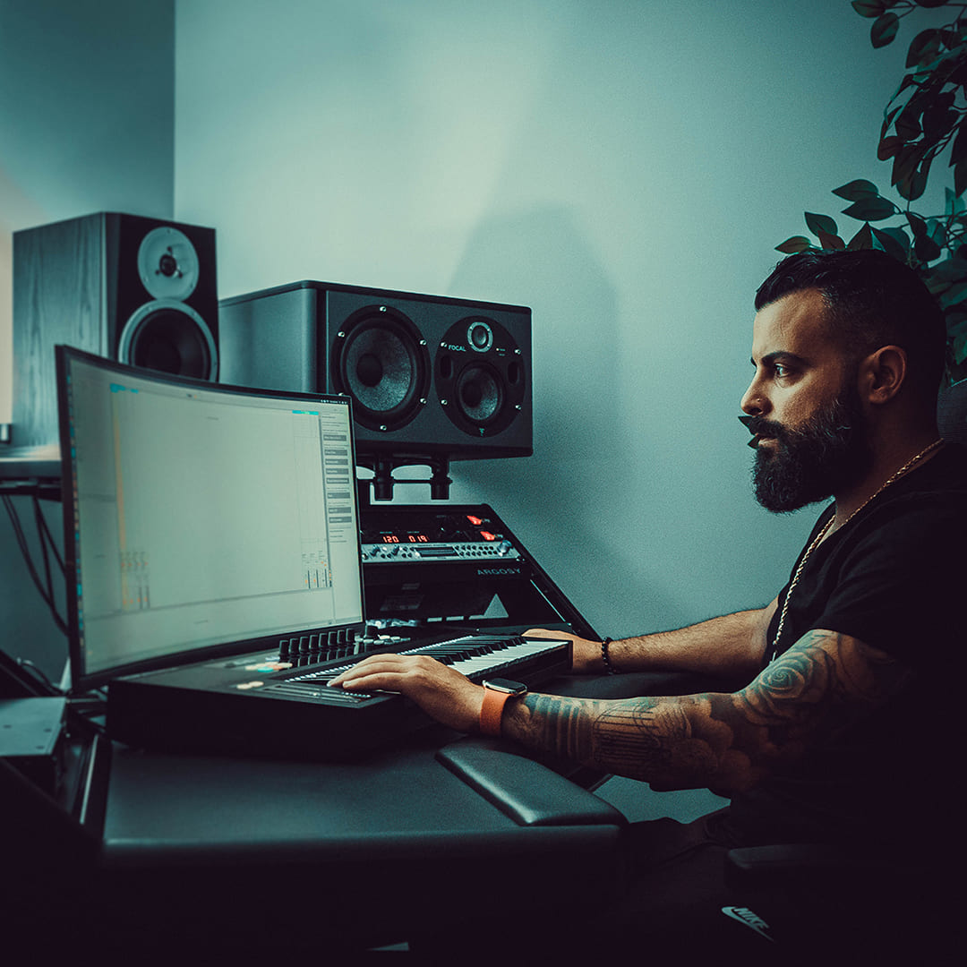 un productor musical trabajando en un estudio, frente a una pantalla y utilizando un teclado MIDI. Al fondo, se pueden ver altavoces de monitoreo y otros equipos de producción, reflejando un ambiente profesional y enfocado en la creación musical.